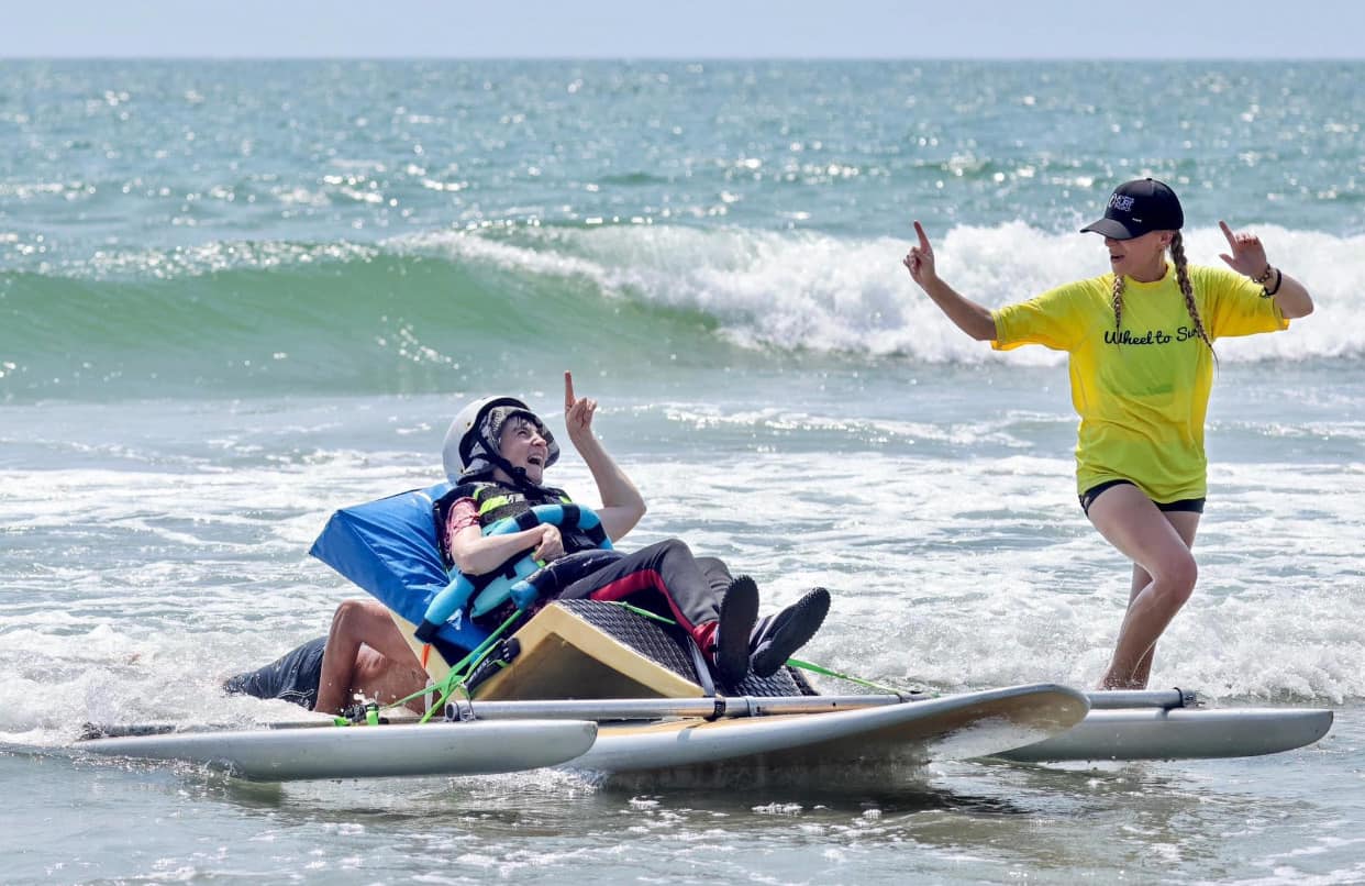 A young person in the water on an adaptive surfboard with a volunteer