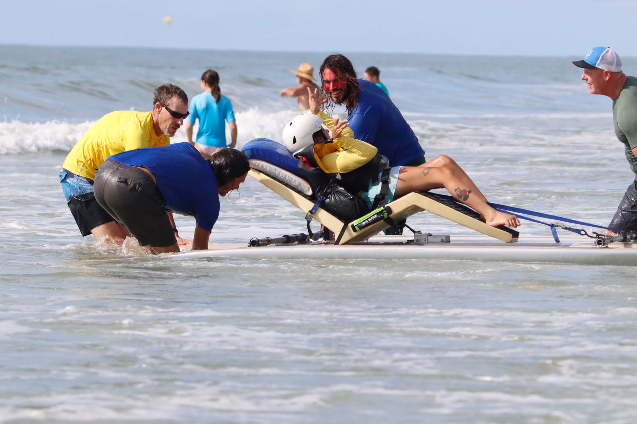 A young person in the water on an adaptive surfboard with a group of volunteers