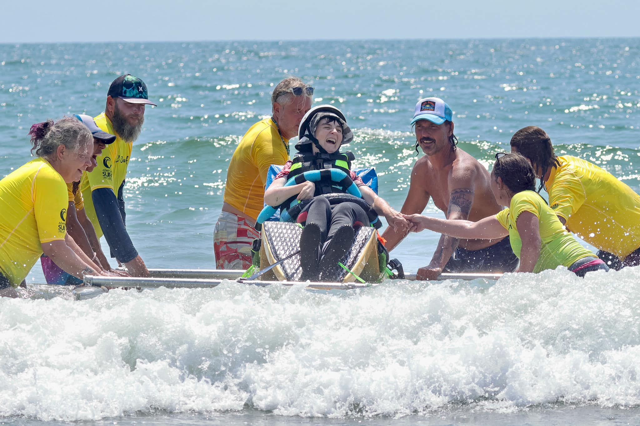 A young person in the water on an adaptive surfboard with a group of volunteers